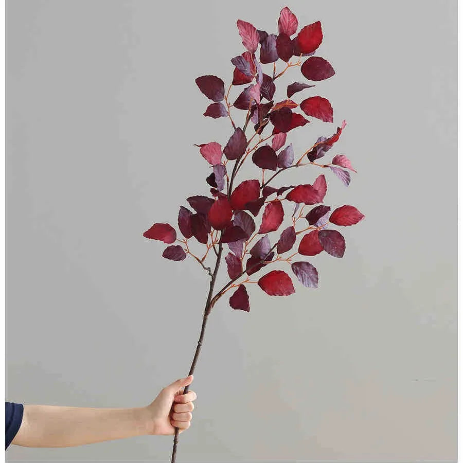 A hand holds a branch of autumn beech simulation flowers with deep red and purple leaves against a gray background.