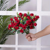 A hand holds a bouquet of red silk roses and green eucalyptus leaves against a white wooden background.