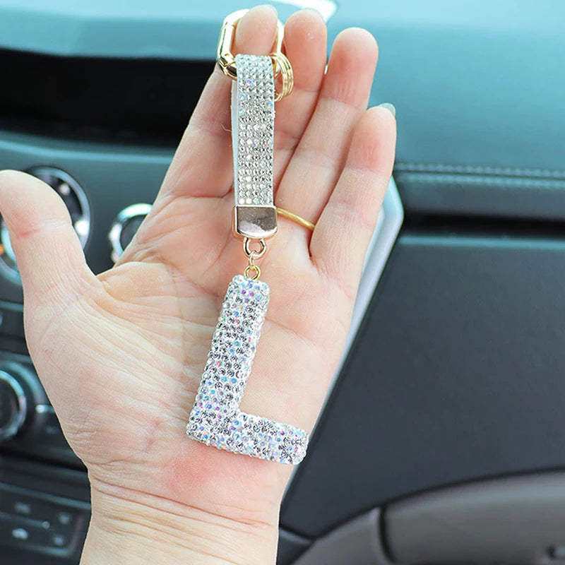A hand holds a crystal letter keychain charm with a gold bail and silver chain.