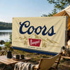 A beige flag banner with dark blue text reading Coors Banquet since 1873 displayed outdoors near a lake.