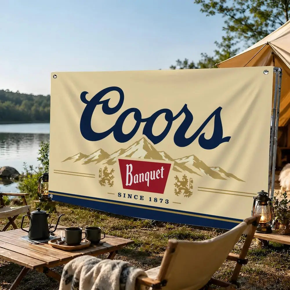 A beige flag banner with dark blue text reading Coors Banquet since 1873 displayed outdoors near a lake.