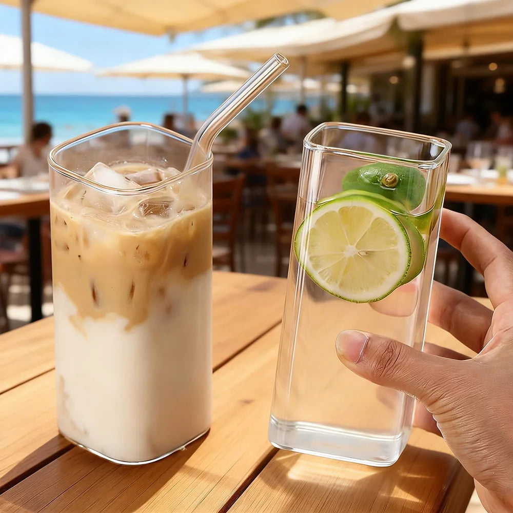 A hand holds a tall glass of iced coffee with milk and ice on a wooden table.