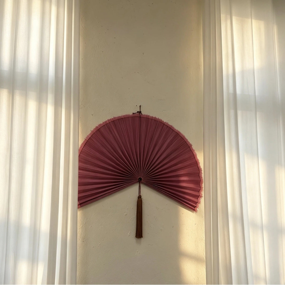 A pink handwoven bamboo wall fan decor with a tassel mounted on a white wall.