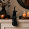 A black witch figurine with a pointed hat and flowing cloak decorates a mantelpiece with pumpkins and candles.