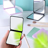 A hand holds an open compact foldable travel makeup mirror with a white surface and black frame.