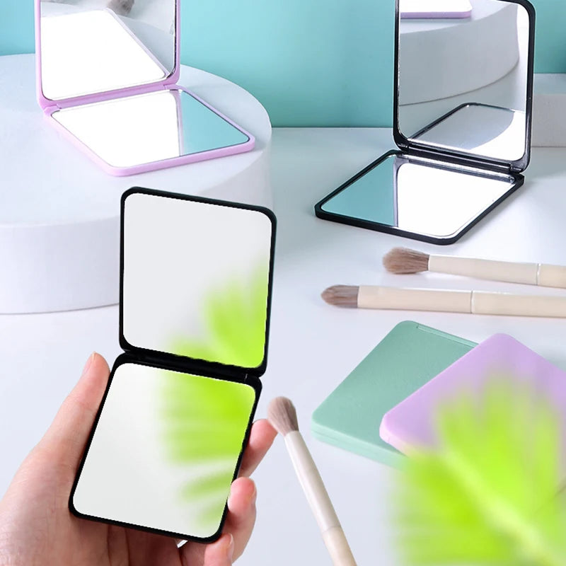 A hand holds an open compact foldable travel makeup mirror with a white surface and black frame.