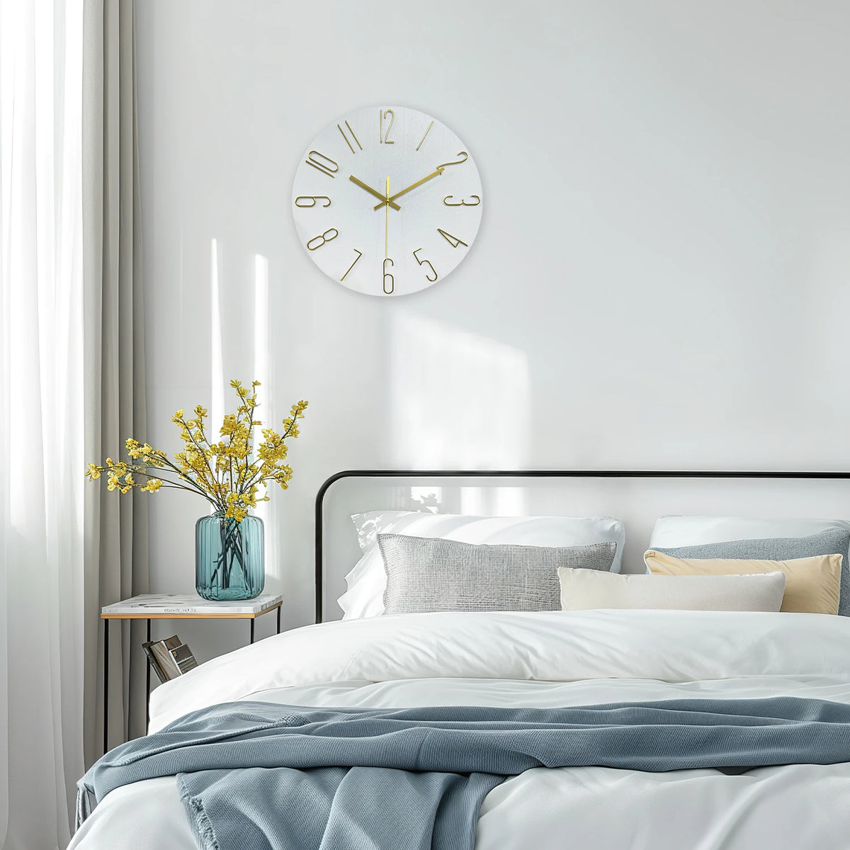 A modern wall clock with gold 3D numerals hangs on a white wall above a bed.