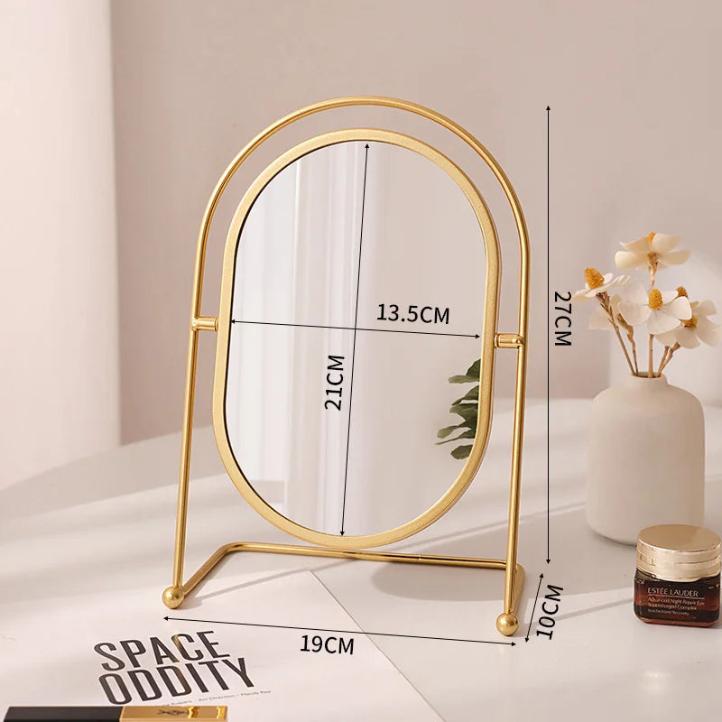 A gold-framed oval vanity mirror with a rotating stand is displayed on a white surface.