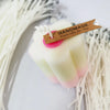 A handmade white candle with pink accents and a wooden tag is displayed against a background of natural cotton fibers.