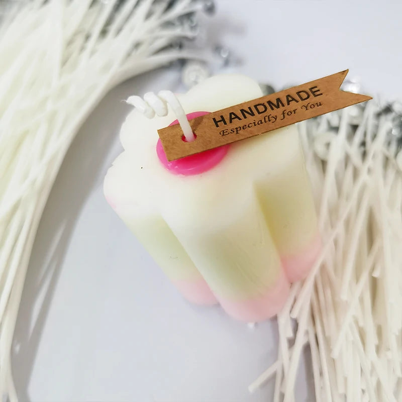 A handmade white candle with pink accents and a wooden tag is displayed against a background of natural cotton fibers.
