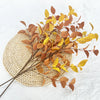 Dried artificial eucalyptus stems in shades of brown and yellow arranged on a woven mat.