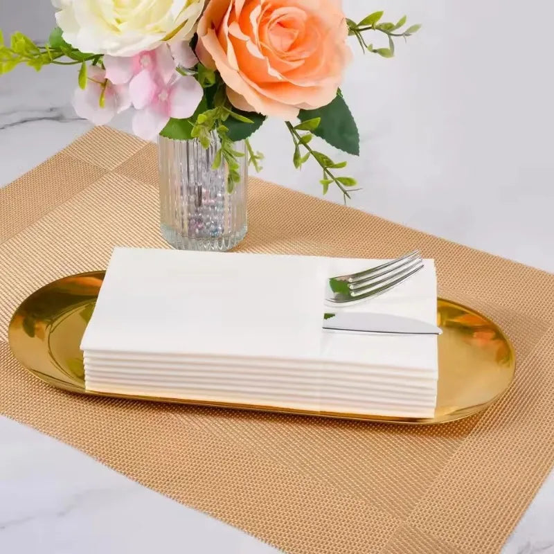 Ein Stapel weißer Einwegservietten auf einem goldenen Tablett mit Blumenarrangements im Hintergrund.