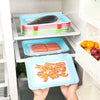 A hand holds a blue reusable food preservation tray filled with cooked shrimp, positioned in a white kitchen storage shelf.