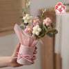 A hand holds a pink paper bouquet featuring white and pink flowers with green leaves.