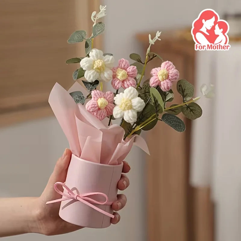 A hand holds a pink paper bouquet featuring white and pink flowers with green leaves.