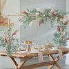 A wooden table displays an adjustable balloon arch stand with gold clamps and greenery for parties.