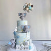 A tiered cake featuring elephant figures and balloons, decorated in light blue and gray tones for a birthday party.