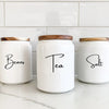 Three white glass canisters with wooden lids labeled Beans, Tea, and Salt sitting on a white countertop.