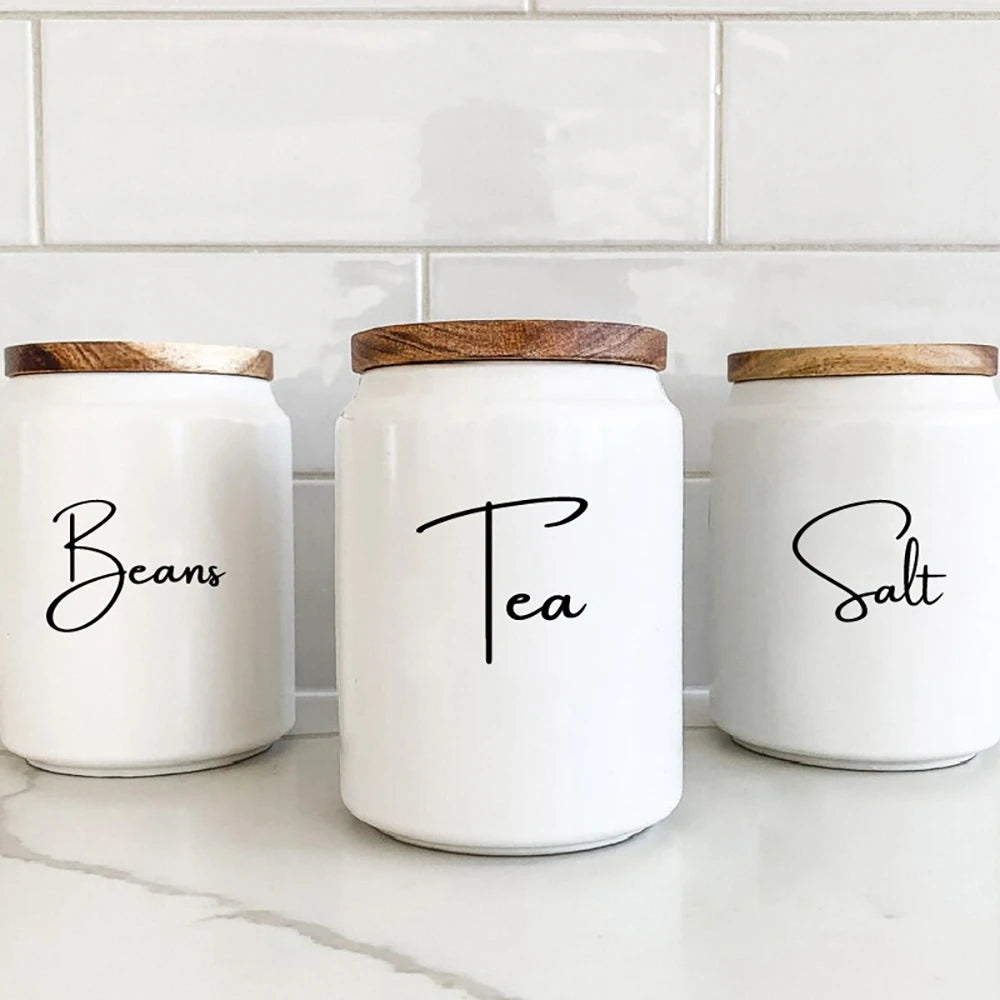 Three white glass canisters with wooden lids labeled Beans, Tea, and Salt sitting on a white countertop.