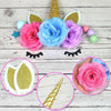 A collection of unicorn cake decorations including pink and blue flowers, gold stars, and colorful balls on a white wooden ba