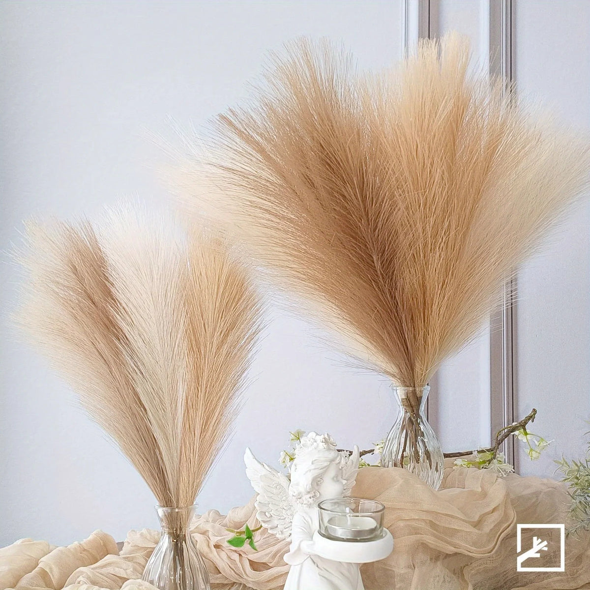 A dried pampas grass floral decor set featuring light beige plumes displayed in glass vases on a draped fabric.