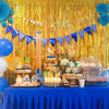 A birthday party setup featuring a gold fringe curtain backdrop, blue and yellow balloons, and decorated treats on a blue tab