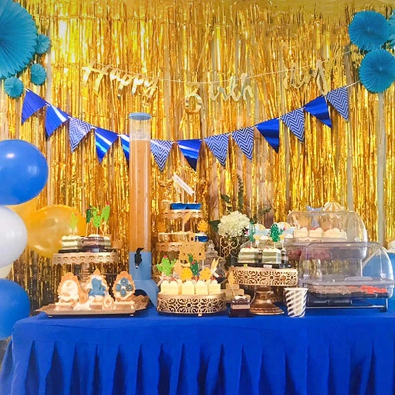 A birthday party setup featuring a gold fringe curtain backdrop, blue and yellow balloons, and decorated treats on a blue tab