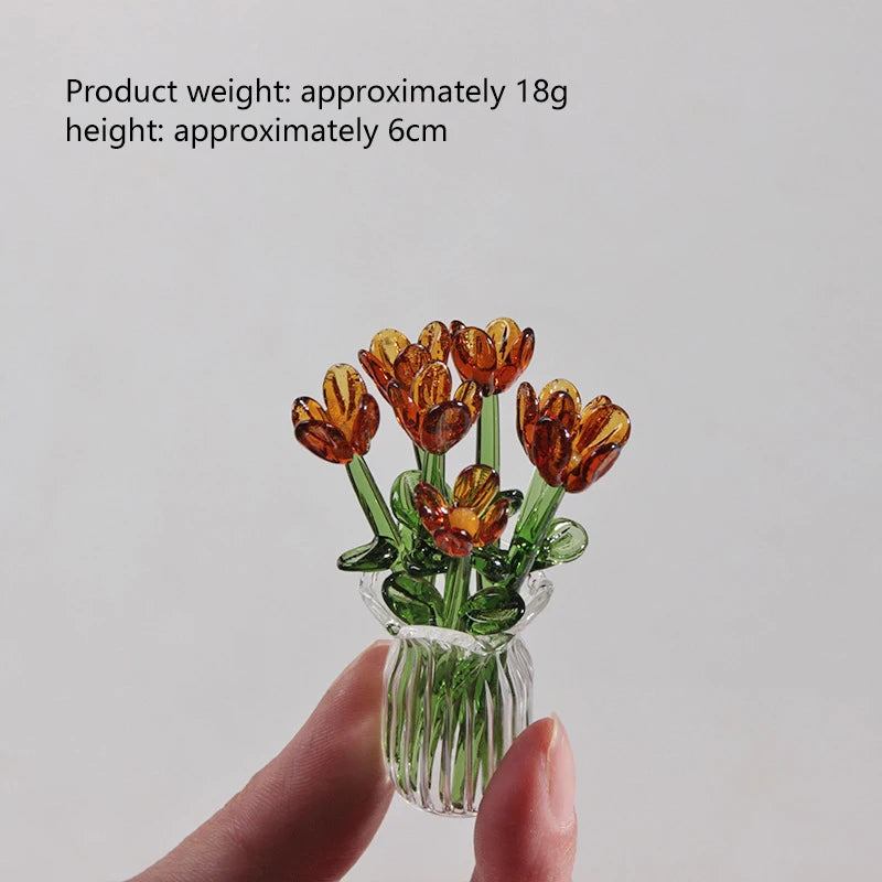 A hand holds a small bouquet of dried orange glass flowers in a clear glass vase.