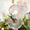 A large, iridescent, transparent number balloon shaped like the number 3 surrounded by green foliage and flowers.
