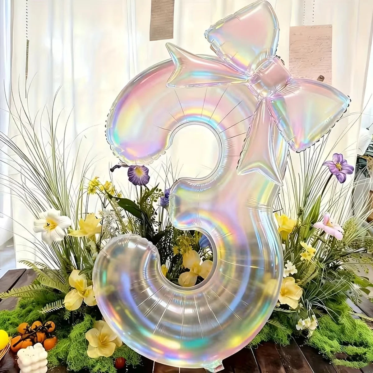 A large, iridescent, transparent number balloon shaped like the number 3 surrounded by green foliage and flowers.