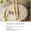 Two long-stemmed printed candles in cream with iris floral designs displayed on a lace platter.