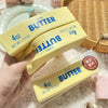 A hand holds a yellow butter squeeze toy labeled 4oz salted butter, resting on a white plate and woven mat.