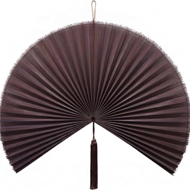 A dark brown handwoven bamboo wall fan decor with radiating slats and a tassel.
