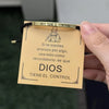 A hand holds a gold-toned bracelet with an engraved design and a tag that reads Dios Tiene El Control