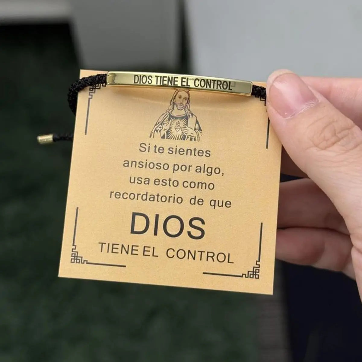 A hand holds a gold-toned bracelet with an engraved design and a tag that reads Dios Tiene El Control