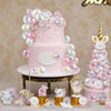 A two-tiered pink cake decorated with clear and pink balloons and floral accents on a gold stand.