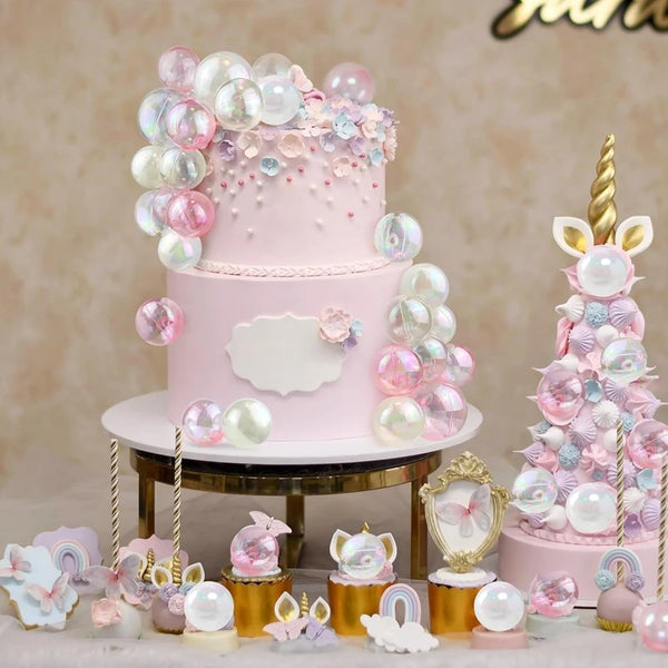 A two-tiered pink cake decorated with clear and pink balloons and floral accents on a gold stand.