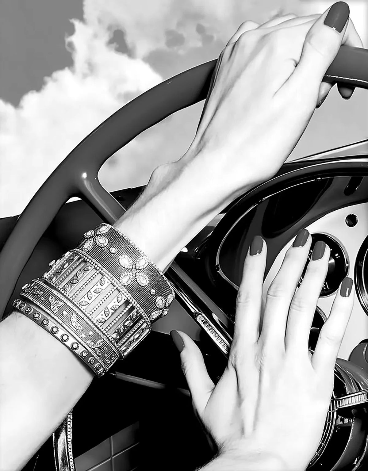 A hand wearing a heavy silver bracelet grips a red steering wheel in a black and white photo.