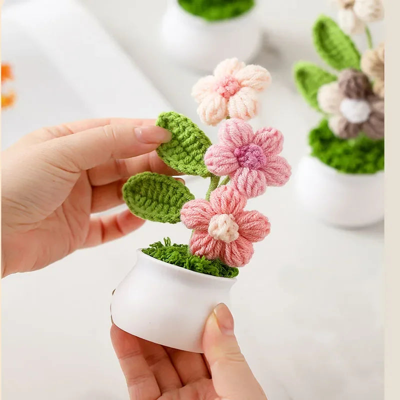 A hand holds a small white pot containing a hand-woven crochet flower plant with pink and green leaves.