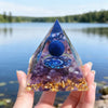 Hand holding a handmade resin orgonite pyramid with purple obsidian and gold copper coil over water