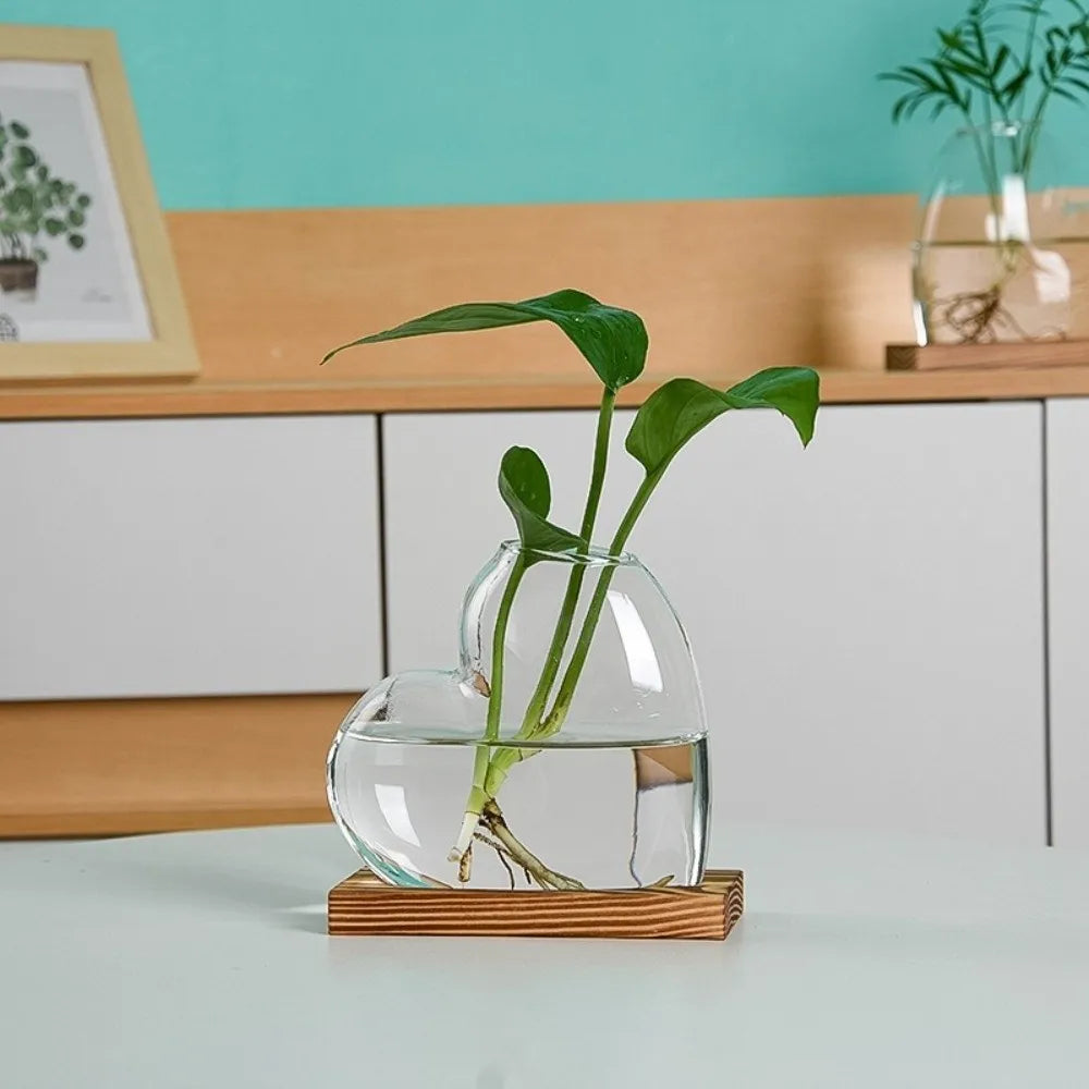 A glass vase shaped like a heart holding a green plant on a wooden stand.