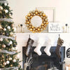 A golden silk Christmas ball wreath ornament displayed on a white mantelpiece next to a decorated Christmas tree.