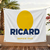A white flag featuring a large yellow circle and the text RICARD DEPUIS 1932, displayed outdoors.