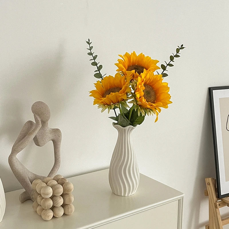 A white ceramic bud vase holds a bouquet of bright yellow sunflowers on a white table.