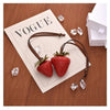 A brown leather strap charm featuring a 3D strawberry and clear ice crystals, displayed on a paper with the word VOGUE.