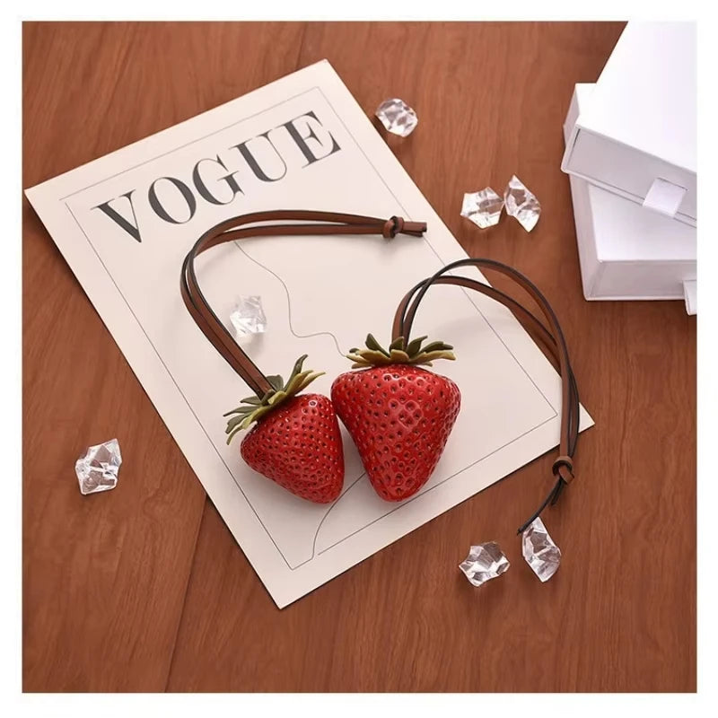 A brown leather strap charm featuring a 3D strawberry and clear ice crystals, displayed on a paper with the word VOGUE.
