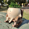 A beige resin ornament shaped like a smiling face with green foliage, sitting in a wooden chair outdoors.