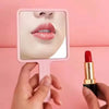 A hand holds a compact handheld makeup mirror with a pink frame, reflecting the user's face.