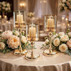 A display of four gold-toned metal pillar candle holders on a table decorated with white and blush roses.