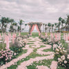 A green aisle decorated with pink rose petals leading to an archway surrounded by greenery and palm trees for a wedding.